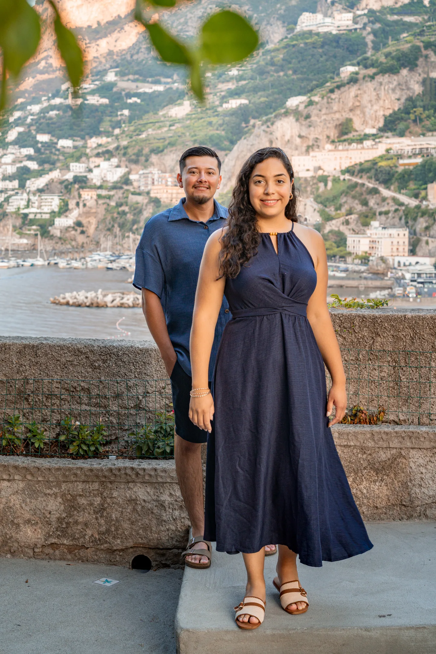 The couple standing beside a low sea wall under the trees